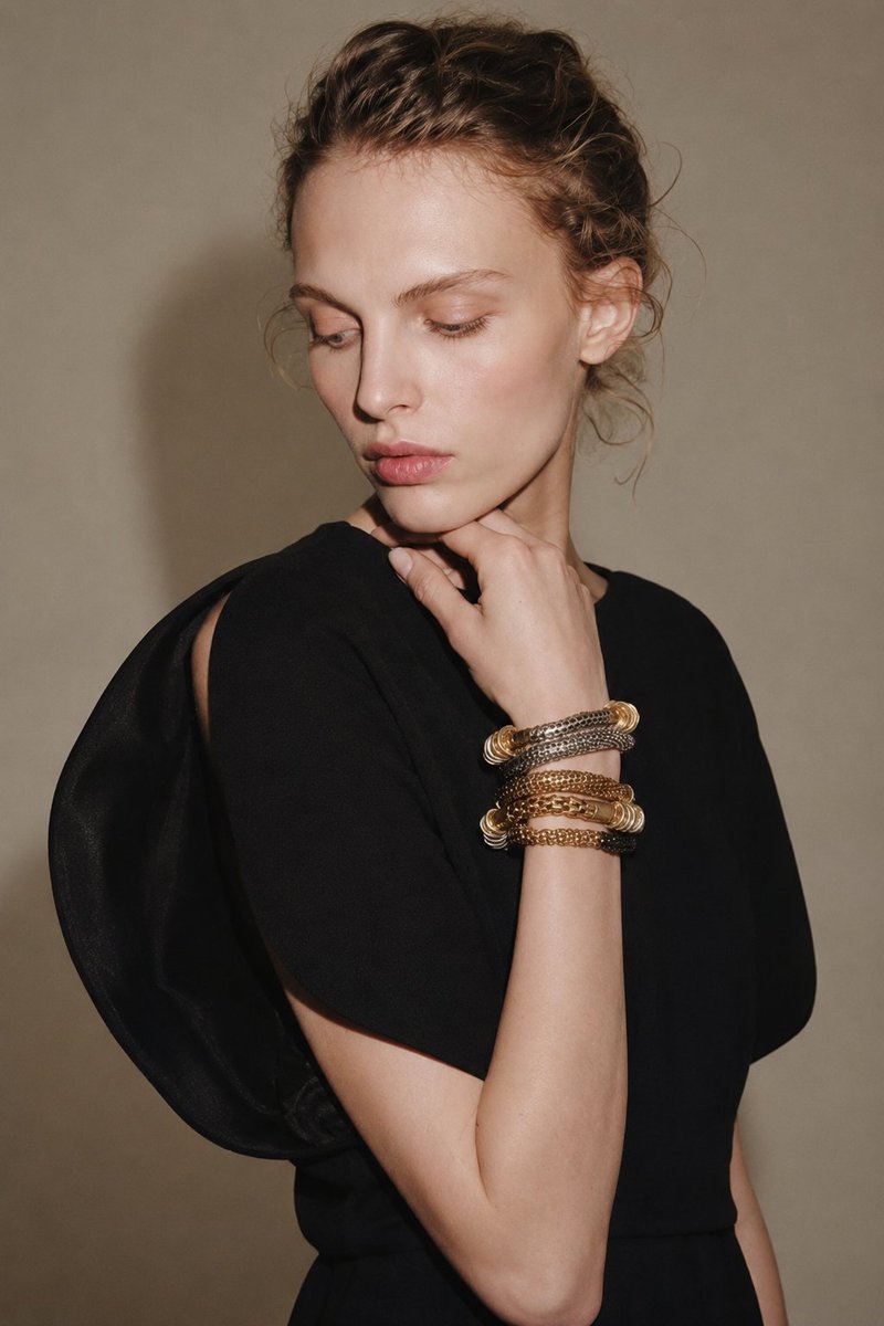 创作一张写实的竖版高级时装摄影棚肖像照，模特年龄21岁以上，背景是一面暖米色墙。画面构图从腰部到头部，模特位于画面中心偏右的位置。她表情优雅，目光看向左侧而非直视镜头，双唇微微张开，涂抹着光泽感十足的唇彩，妆容自然清淡，眉形精致，眼影是淡淡的蜜桃色，肌肤散发着水润光泽，颧骨处进行了高光修饰。

她的头发是浅棕色到深金色，梳成一个略带湿润感的松散发髻，鬓角、耳后和颈后留有几缕卷曲的碎发。整体造型应简洁、现代，并带有T台风情，展现自然的纹理和些许碎发。

她身着一件极具戏剧张力的黑色雕塑感上衣或连衣裙，面料为极简的哑光质感。这件服装拥有不对称的建筑感廓形：一侧是流畅圆润的肩部拼接，上臂处有镂空设计，简洁的高领口，以及柔软的褶皱在肩部和上半身营造出丰盈感。黑色面料应与她温暖的肤色和中性背景形成鲜明对比。

她的姿态优雅而沉思：一只手臂交叉于腰间，另一只手抬至下巴，手指放松地轻触下颌线。抬起的手腕上佩戴着层叠的奢华手镯，材质混搭，包括金色、银色和青铜色的链条、盘绕的手镯、抛光的戒指以及编织金属饰品。这些手镯应精致闪亮，成为脸部附近的视觉焦点。

背景简洁明快：光滑的米色墙面，在模特身后投射出柔和的阴影。灯光应温暖、定向且富有时尚感，从左前方照射，在模特的脸部、额头、嘴唇、手镯、手、手臂和黑色服装上营造出强烈而又不失美感的亮点。在墙面和雕塑般的肩部轮廓下方添加柔和的阴影。运用逼真的肌肤纹理、清晰的珠宝细节、干净利落的布料边缘以及微妙的高级时装对比。

风格：写实奢华时尚大片摄影，成人模特，黑色雕塑感设计师上衣，叠戴金属手镯，光滑的湿发造型，暖米色摄影棚墙面，沉思的侧视，戏剧性的定向灯光，精致的T台氛围，逼真的肌肤纹理，清晰的珠宝细节，竖幅比例，高细节，无文字，无水印，无标志，无其他人物，无变形人体，无卡通风格，无过度曝光，无杂乱元素。